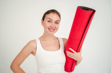 Hevesli bir kadın, elinde yoga minderini tutarken gülümseyerek canlandırıcı bir egzersiz seansına hazırlanıyor. Heyecanını, zindeliğini ve refahını parlak ve neşeli bir şekilde gösteriyor. Yüksek