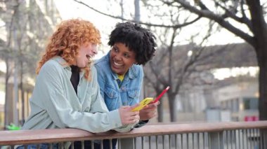 Two cheerful young women enjoying their time together as they engage with their smartphones, sharing laughter and joy while using mobile applications in a lively atmosphere.