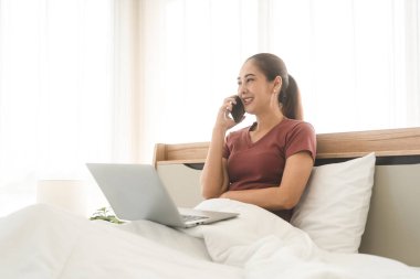 Lifestyle routine with internet technology concept. Young adult asian woman using laptop and talk with smartphone on bed for telemedicine mental health. People rest in bedroom activity.