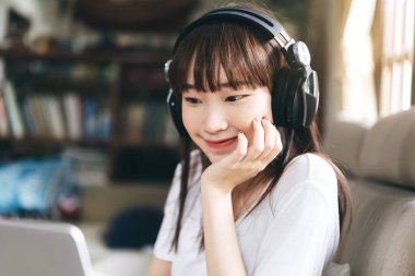Relax and stay at home for new normal concept. Happy asian teenager woman listen music via internet streaming with headphone. Living room background with window light.