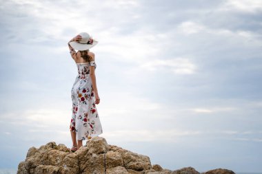 Adult asia woman travel to the sea and standing relax on the rock looking to sky. Hand holding the hat in dress cloth with rear view. Pattaya, Thailand.