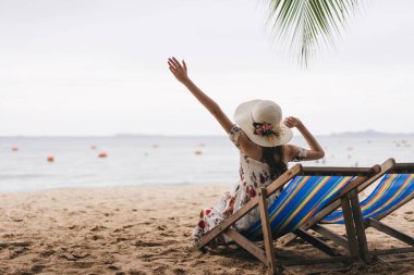 Summer beach holiday concept. Young asian travel woman relax on beach chair hand up her hand with floppy hat. Sand cler sky background with copy space. Pattaya, Thailand
