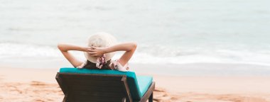 Summer beach holiday concept. Young asian healthy traveling woman relax on beach chair arm up her hand with floppy hat. White sand and sea background banner with copy space. Pattaya, Thailand