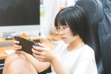 İnek stili genç Asyalı oyuncu kadın gözlük takıyor ve el bilgisayarında oyun oynuyor. Zafer havası için rekabet. İnsanlar evlerinde boş zaman geçiriyorlar..