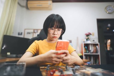 People lifestyle with interesting hobby at home concept. Young adult asian woman solo playing board game on top table. Using mobile phone for online application multiplayer