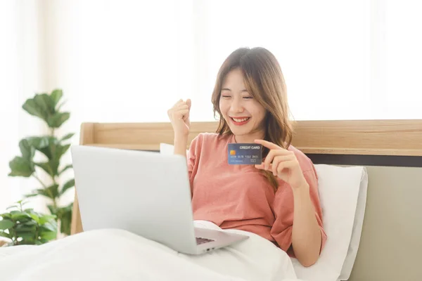 People lifestyle in bedroom after wake up in morning concept. Young adult single asian woman using laptop computer and credit card for shopping online.