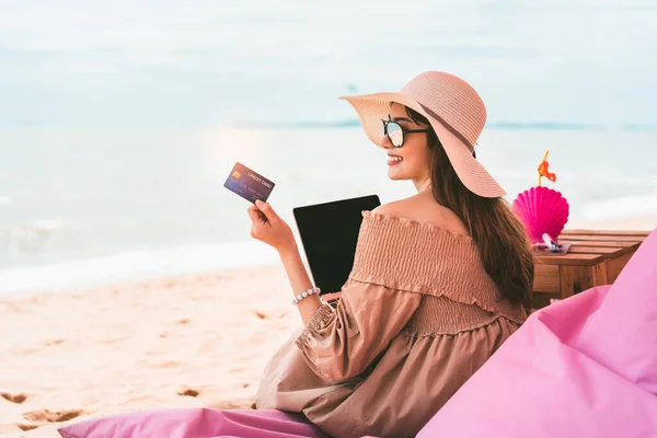 Beautiful asian smile woman relax at beach on summer vacation. Holding credit card with computer. Digital payment concept for shop online and booking hotel when travel. Pattaya, Thailand.