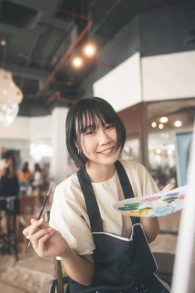 Portrait of happy young adult asian woman painting brush on canvas at workshop art lesson class. People leisure with creativity education lifestyle for mental health concept.