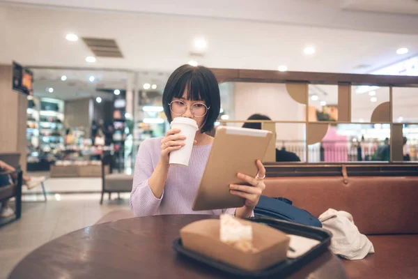Young adult business asian woman sipping coffee and using tablet for online internet at indoor public cafe on day. Single people lifestyle in city concept.