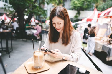 Happy young asian adult woman casucal city lifestyle on weekend at cafe. Relax at restaurant using smart phone for play game wood table with coffee and tablet. Outdoor area on day time.