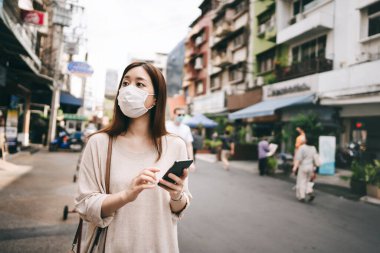 Casual business young asian single woman wear mask for new normal lifestyle protect corona virus or covid19. Using smartphone on sidewalk street outdoor area on day. Bangkok, Thailand.