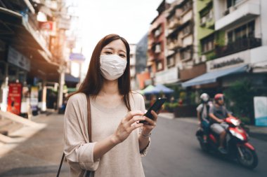Casual business young asian single woman wear face mask for new normal lifestyle protect corona virus or COVID-19. Using smartphone on sidewalk street outdoor area on day. Bangkok, Thailand.