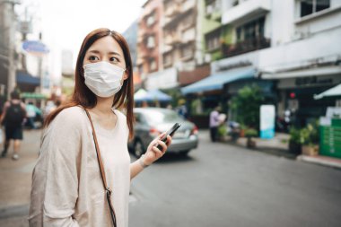 Casual business young asian single woman wear face mask for new normal lifestyle protect corona virus or COVID-19. Using smartphone on sidewalk street outdoor area on day. Bangkok, Thailand.
