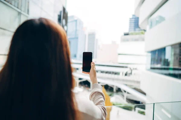 Shoulder view of woman holding mobile phone empty screen with blur city background on day.