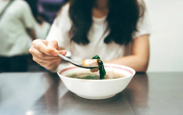 Poeple travel and eating street food concept. Chinese style crispy pork soup on spoon. Woman blur background at sidewalk restaurant. Chinatown Yaowarat market, Bangkok, Thailand.