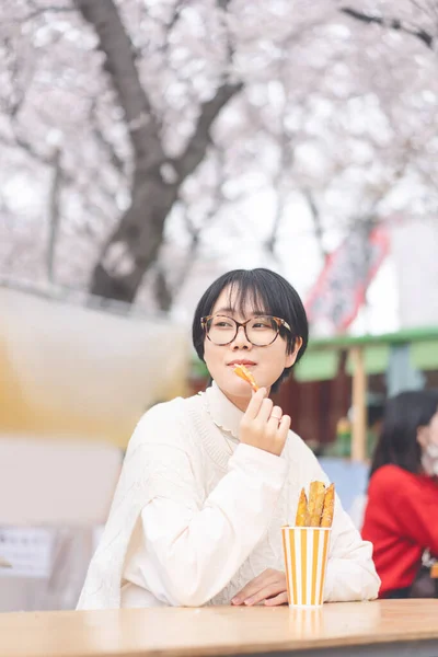 縦の肖像若い大人のアジアの観光客の女性が座って 日本のスタイルの通りの食べ物を食べる 桜の木春祭りで日本旅行シーズン — ストック写真