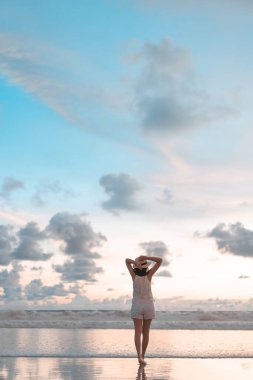 Genç yetişkin gezgin Asyalı kadının dikey görüntüsü kumsalda dinleniyor ve dramatik bir gün batımı gökyüzü var. Yaz tatillerinde seyahat..