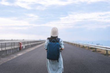 Rear view of young woman traveller with blue backpack walking on road. People solo outdoor travel on summer holidays concept. Background with blue sky. clipart