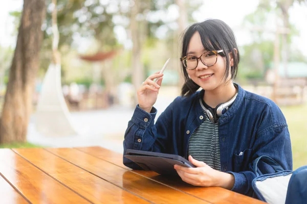 フリーランスのデジタルノマドワークと研究の人々の概念 若い大人のアジアの女性は タブレットを使用して眼鏡やヘッドフォンを着用します 屋外の木製のテーブルに座る 青いテーマカラーの衣装 — ストック写真