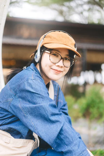 アウトドアライフスタイルのコンセプトで人々の精神療法 若いアジアの女性は帽子のデニムジャケットと眼鏡を身に着けているファッションヘッドフォンで音楽を聞くリラックス 縦の肖像 — ストック写真