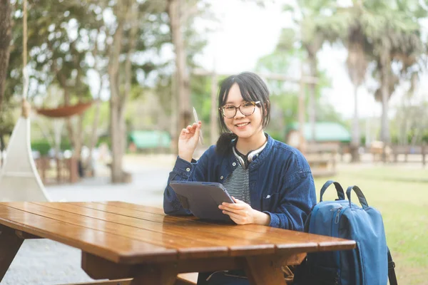 フリーランスのデジタルノマドワークと研究の人々の概念 若い大人のアジアの女性は タブレットを使用して眼鏡やヘッドフォンを着用します 屋外の木製のテーブルに座る 青いテーマカラーの衣装 — ストック写真