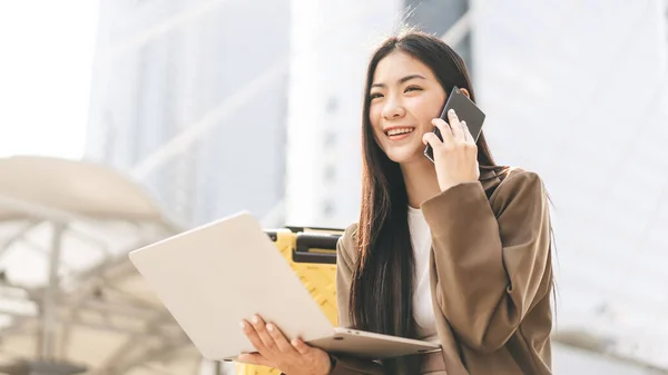 商业自由职业的亚洲成年女性使用笔记本电脑和智能手机 美丽的微笑面对长发 每天都带着旅行箱坐在户外 城市居民的生活方式 — 图库照片