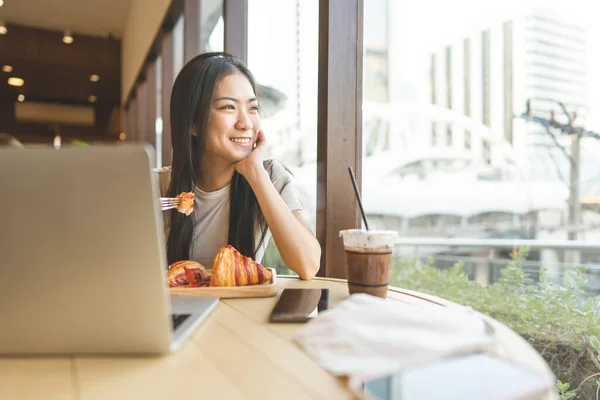 人们在咖啡馆休息的概念 年轻的亚洲女人吃面包时笑容满面 桌上有手提电脑 白天坐在室内靠窗的位置 — 图库照片