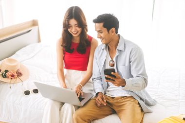 İnsanoğluna odaklan. Mutlu bir gülümseme genç yetişkin güneydoğu Asyalı çift tatil gezisi için dizüstü bilgisayar kullanıyor. Evde yaşıyor, şapka ve bavullarla yatakta oturuyor..