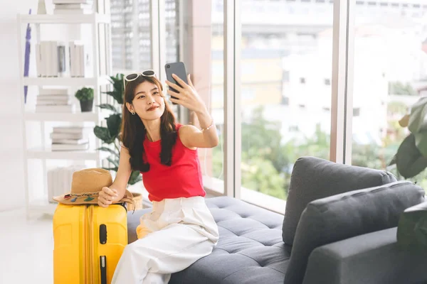 黄色の旅行荷物を持つ若い大人の東南アジアの女性の肖像画 旅を開始する前に自撮りのためのスマートフォンを使用 家のコンセプトで旅行の準備をする — ストック写真