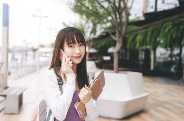 Üniversiteli insanların gündüz konsepti. Cep telefonu kullanan mutlu, genç, Asyalı bir kadının portresi. Not defteri kırtasiyesini tut ve açık havada sırt çantası tak.