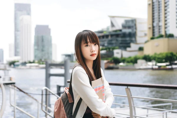 大学の人々の生活様式の日の時間概念 若い大人のアジアの学生女性の肖像画 ノートを持ち 屋外でバックパックを着用してください 都市と川のぼかしの背景 — ストック写真