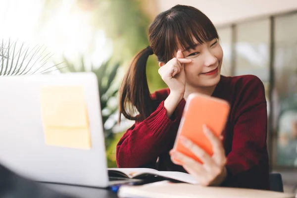 快乐的亚洲青年女学生穿着红色毛衣 使用手机和笔记本电脑在咖啡店上网学习 大学生的生活方式与现代技术日新月异 — 图库照片