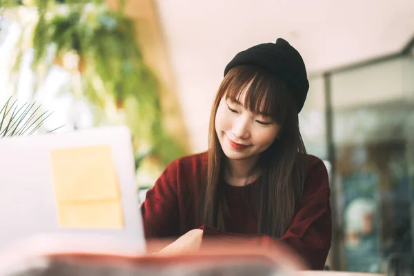 年轻的亚裔女大学生穿着红色毛衣 头戴羊毛帽 使用笔记本电脑学习 并在咖啡店上网工作 大学生的城市生活方式则是在冬日出门在外 — 图库照片