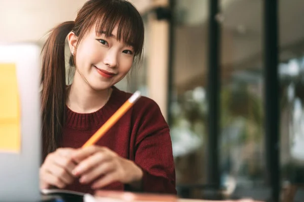 年轻的亚裔女大学生穿着红色毛衣 使用笔记本电脑学习 并在咖啡店上网工作 大学生的城市生活方式则是在冬日出门在外 — 图库照片