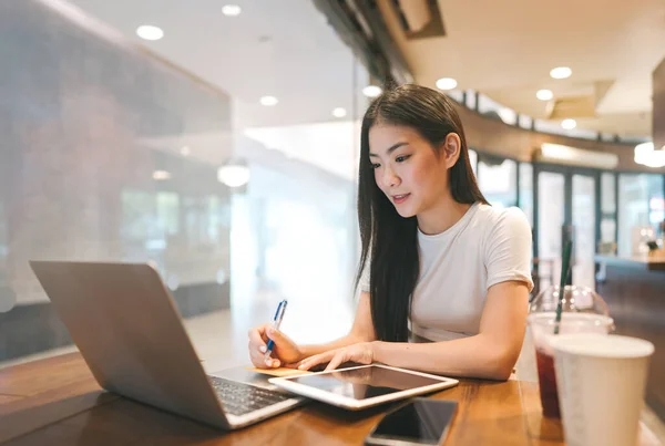 商业自由职业的亚洲年轻女性 使用笔记本电脑 在咖啡厅桌上写便条 城市人的生活方式与现代技术日新月异 数字游牧民 — 图库照片