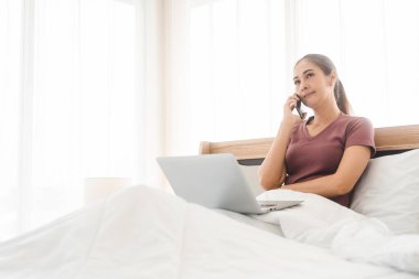 İnternet teknolojisi konseptiyle yaşam tarzı rutini. Genç Asyalı kadın diz üstü bilgisayar kullanıyor ve teletıp akıl sağlığı için yatağında akıllı telefonla konuşuyor. İnsanlar yatak odasında dinleniyor..
