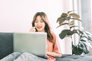 Güneydoğu Asyalı genç bir kadın kanepede dizüstü bilgisayarla evde kalıp çalışıyor. İnternet teknolojisiyle insanların yaşam tarzı. Gün arkaplanındaki pencere ışığı. 