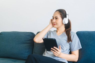 İnsanlar ev kavramında mutlu bir yaşam tarzına sahipler. Genç Asyalı kadın kulaklık takıyor müzik dinliyor ve dijital tablet kullanarak rahatlıyor. Mavi kanepede otururken.