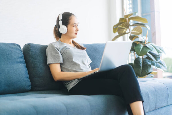 People happy lifestyle satisfation at home concept. Young adult asian woman wear headphone listen music and relax using laptop computer. Sitting on blue sofa.