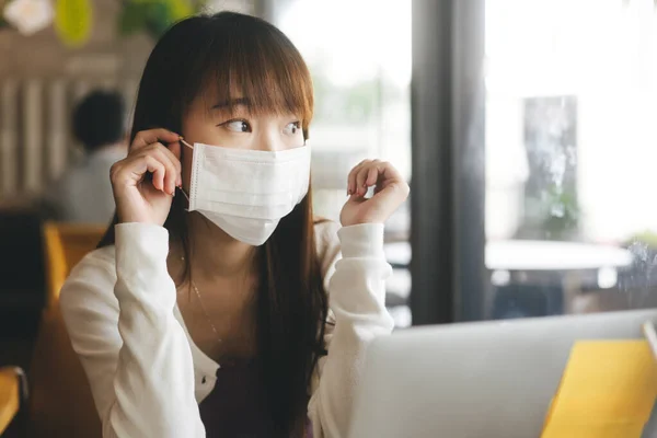 Young adult asian female with protective face mask for virus corona or covid 19. Work and study at cafe new normal during pandemic lifestyle concept. Indoor background on day