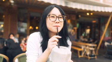 Tayland restoranında gözlüklü, hindistan cevizi suyu içen Asyalı genç bir kadın. Şehir kırılma kavramında insanlar rahatlar.