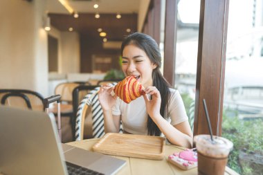 İnsanlar kafe konseptinde rahatlıyor. Genç Asyalı bir kadının kruvasan ekmeği yediği mutlu bir gülümseme. Laptop bilgisayarıyla masada. Gündüzleri pencerenin yanında oturmak.