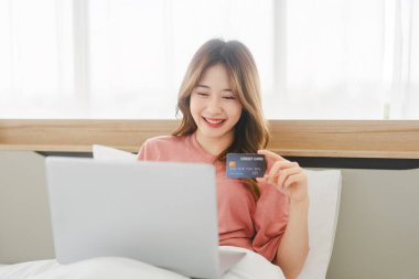 People lifestyle in bedroom after wake up in morning concept. Young adult single asian woman using laptop computer and credit card for shopping online.