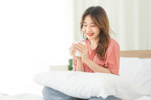 Serenity of a young Asian woman holding a coffee cup. Enjoy the peaceful morning time as she sits on a white bed, with happy smile. Moment of relaxation and contentment.