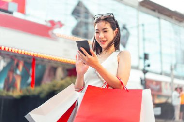 İnsanlar şehir yaşam tarzlarını alışveriş tüketimiyle satın alıyorlar. Genç Asyalı kadın dijital cüzdan bankacılığı için akıllı telefon kullanıyor. Mutlu gülen surat dışarıda duruyor..