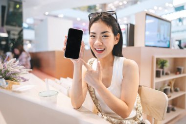 İnsanlar serbest dijital göçebe işi konsepti uyguluyorlar. Mutlu bir gülümseme, genç Asyalı kadın kapalı bir kafede oturuyor. Akıllı telefonun boş ekranını gösteriyor. Etkilenen gen z modern yaşam tarzları.