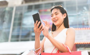İnsanlar şehir yaşam tarzlarını alışveriş tüketimiyle satın alıyorlar. Genç Asyalı kadın dijital cüzdan bankacılığı için akıllı telefon kullanıyor. Mutlu gülen surat dışarıda duruyor..