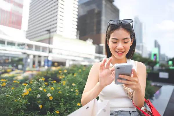 People city lifestyles with buying shopping consumerism. Young adult asian woman using smartphone typing message. Digital wallet banking payment. Happy face sitting at outdoor.