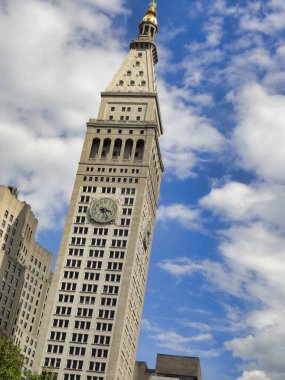 Üzerinde saat kulesi olan bir gökdelen mavi gökyüzüne doğru uzanıyor. Binaların ön cephesi, New York 'taki bir apartman bloğuna benzeyen şehir zeminine karşı dik duruyor.