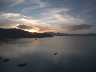 Palawan güneşi dağların ardında batarken, sakin Port Barton Filipinleri 'nin okyanus suyuna yansıyan, canlı renklerle resmedilmiş bir alacakaranlık. Kümülüs bulutları kararmış gökyüzünde dağılır.